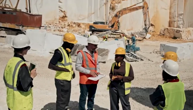 A construction supervisor holding a drawing and explaining plans to a group of five workers, with an excavator in the background, showcasing teamwork and leadership on a construction site.