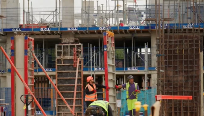 Formworkers on a construction site holding formwork panel hooked to a crane, with concrete columns and rebar in the background, demonstrating the structural work in progress.