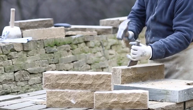 A skilled stonemason chiselling a stone with a hammer and chisel, demonstrating craftsmanship in front of a stone wall.