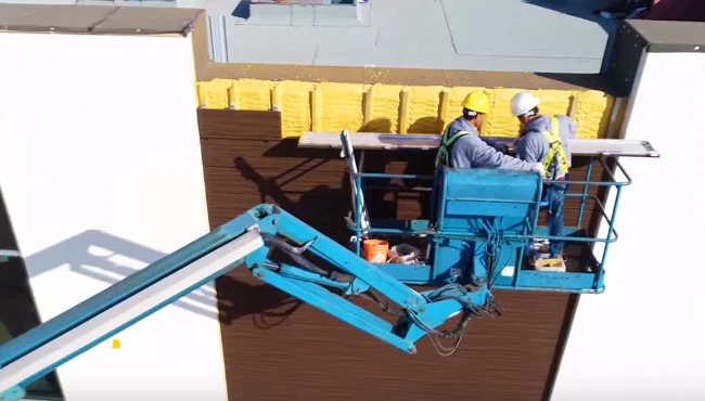 Two skilled cladding installers on a mobile elevated platform, installing a cladding panel on a building structure, demonstrating expertise in exterior building cladding.