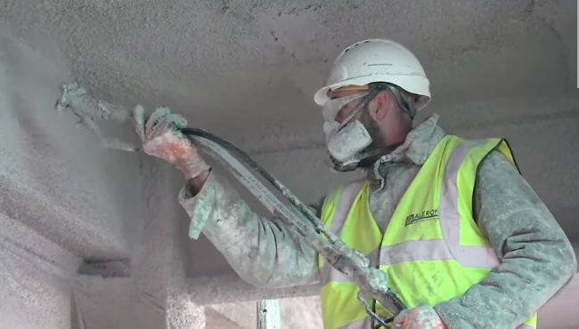 A fire protection installer spraying a wall with fireproof coating, wearing full PPE, to ensure fire safety and compliance in construction.