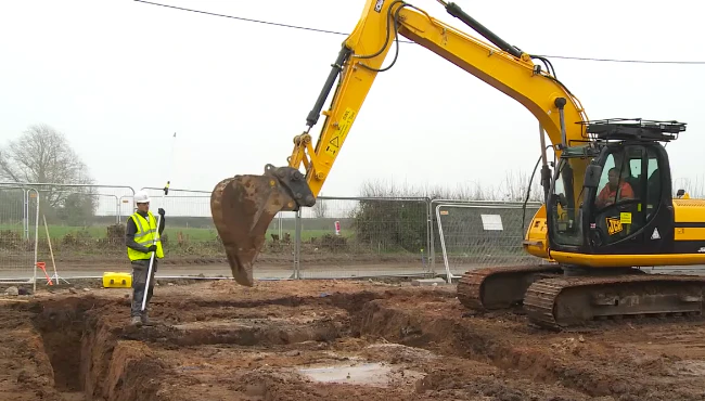 A young groundworker in PPE holding a pole, standing near a yellow excavator and trenches prepared for building foundations on a construction site.