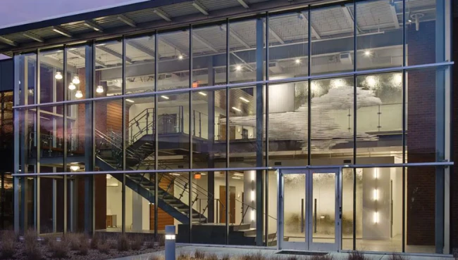A building facade made of glass and aluminum curtain wall, glowing from the inside as evening lighting illuminates the structure.
