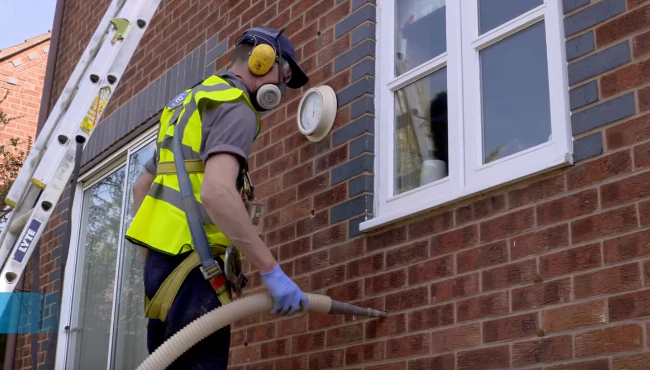 A cavity wall installer wearing PPE blowing insulation into a house cavity using specialized equipment to improve thermal efficiency.