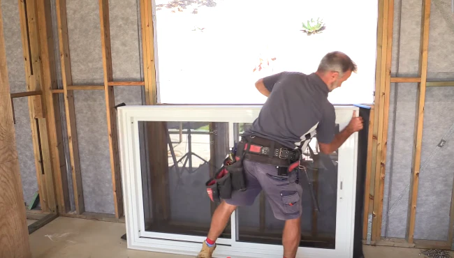 A window fitter on a building site, seen from behind, prepares a window for installation with their hands on the frame, demonstrating focus and expertise.