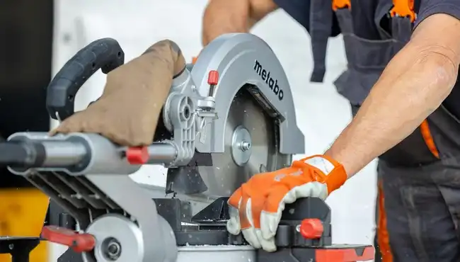 Close-up of a maintenance operative’s hands in orange gloves using a chop saw, demonstrating precision and safety during cutting work.